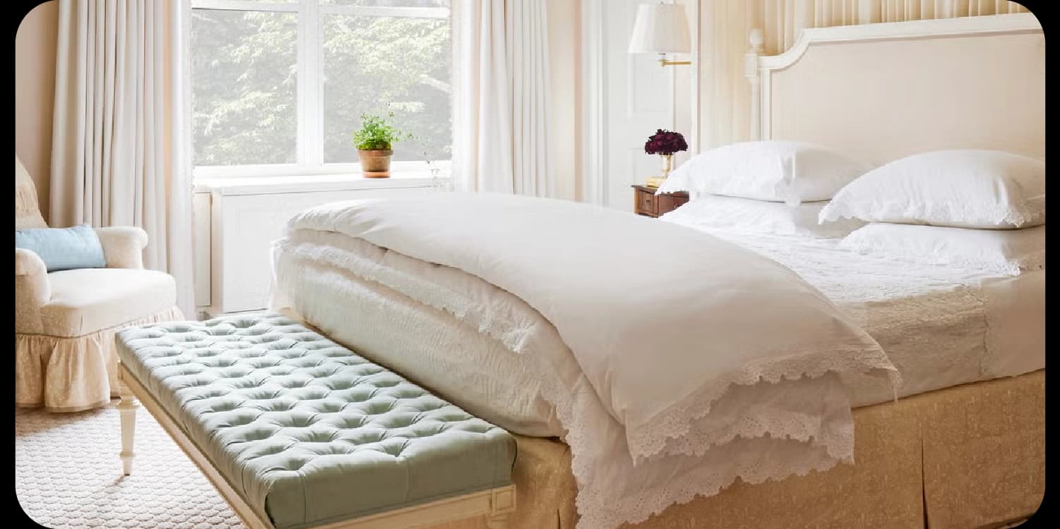 A bedroom with a white bed, a tufted bench, and a window