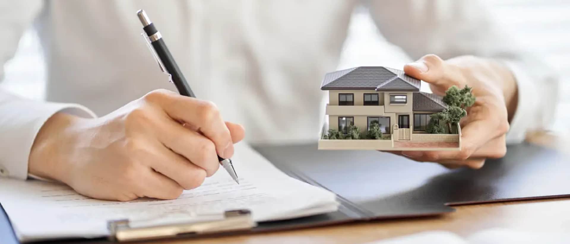 Person signing paperwork while holding a model house